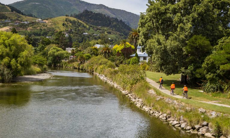 Green summer landscape with three recreational cyclists in orange clothes alongside the river in Nelson, South Island, New Zealand